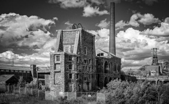 Black and white photograph of the dilapidated, towering stone winding engine house and tall brick chimney stack of Chatterley Whitfield Colliery, Staffordshire, under a cloudy sky.