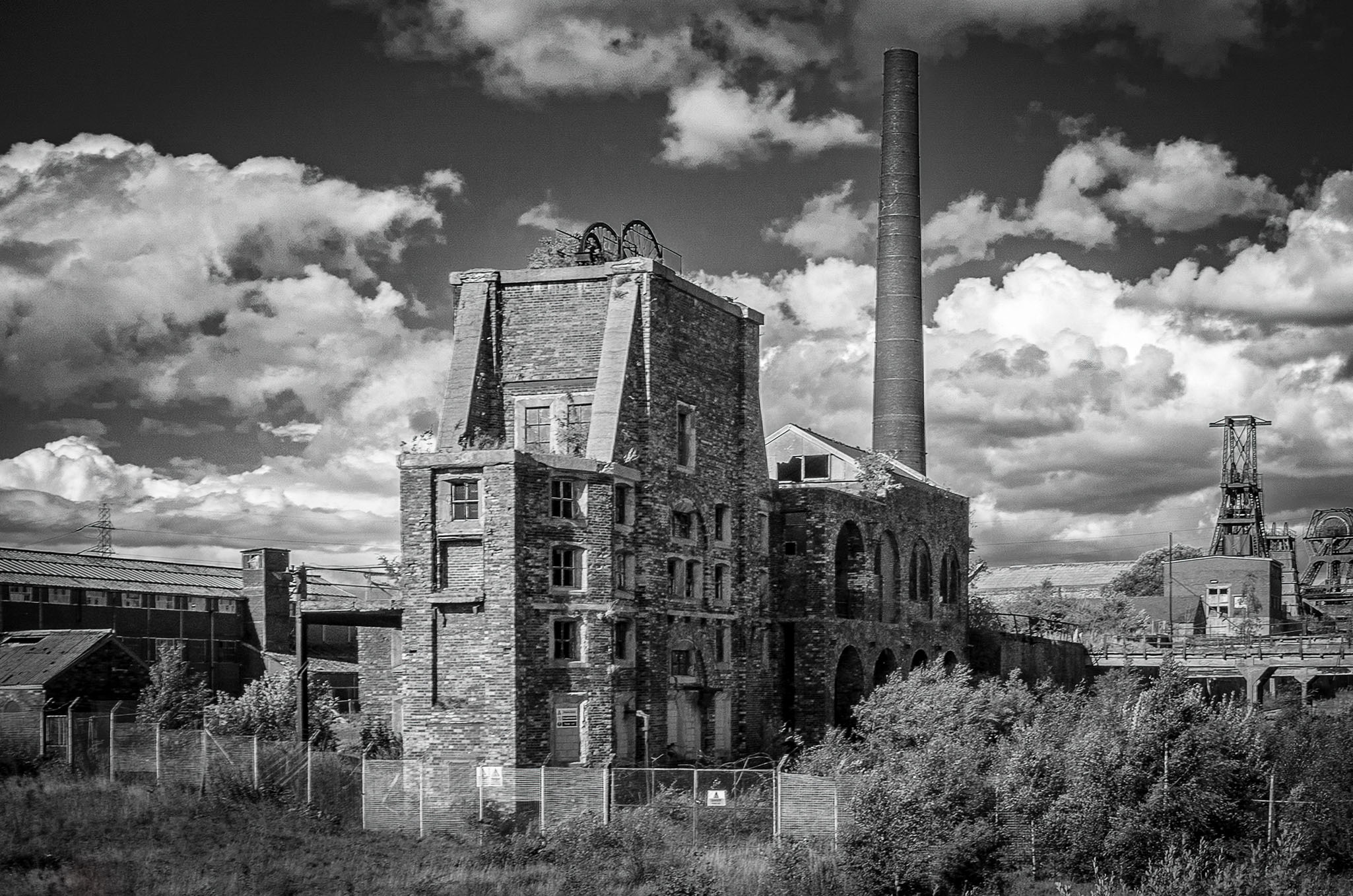 Black and white photograph of the dilapidated, towering stone winding engine house and tall brick chimney stack of Chatterley Whitfield Colliery, Staffordshire, under a cloudy sky.