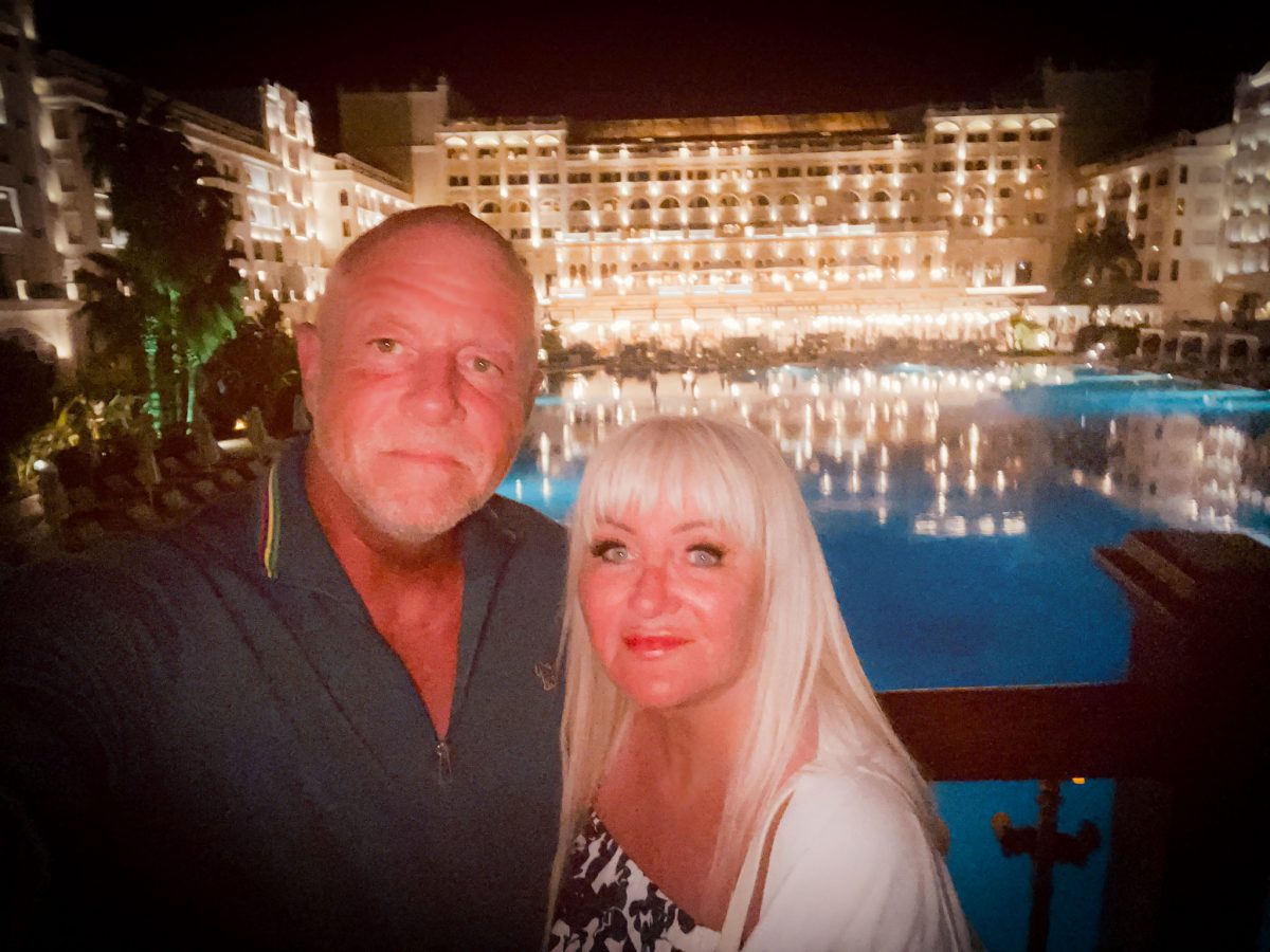 A couple posing for a selfie at night with the illuminated Titanic Mardan Palace hotel and pool in Antalya, Turkey in the background