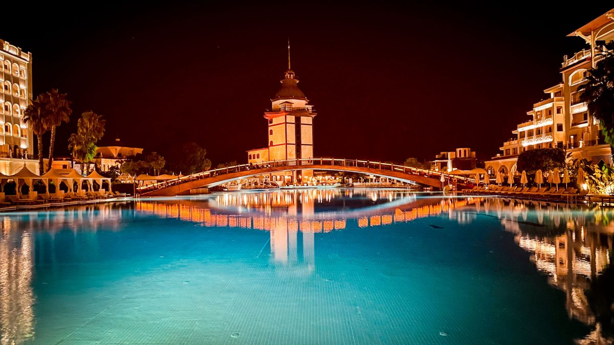 Night view of the illuminated bridge and central tower at Titanic Mardan Palace reflected across the large pool in Antalya, Turkey