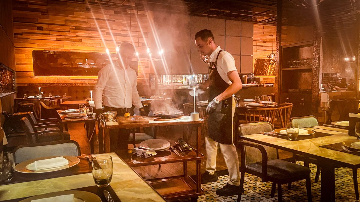 A chef preparing a sizzling dish tableside with steam rising, inside a warm, stylish restaurant at Titanic Mardan Palace in Antalya, Turkey