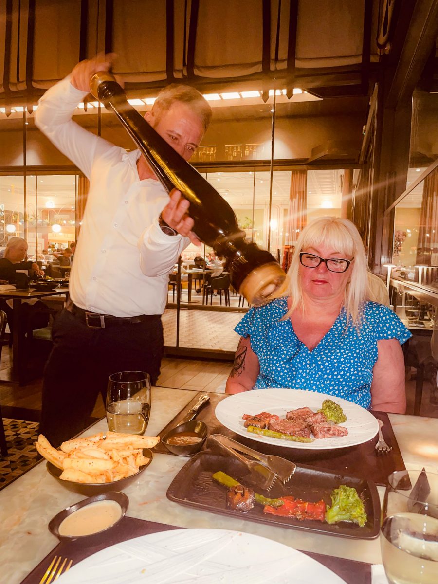 A restaurant server using an oversized pepper grinder to season a diner's meal at a table in Titanic Mardan Palace, with plates of steak, vegetables and fries on the table