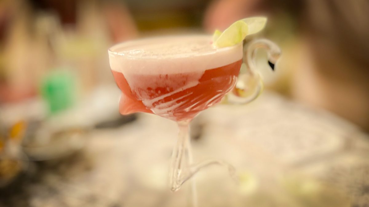 A pink frothy cocktail served in a decorative swan-shaped glass on a patterned table.