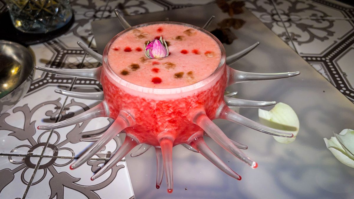 A vibrant red cocktail served in a unique spiked glass with decorative drops and a small rose bud on top.