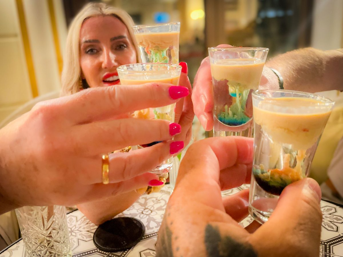 Friends raising colourful layered shot glasses in a celebratory toast at a bar table.