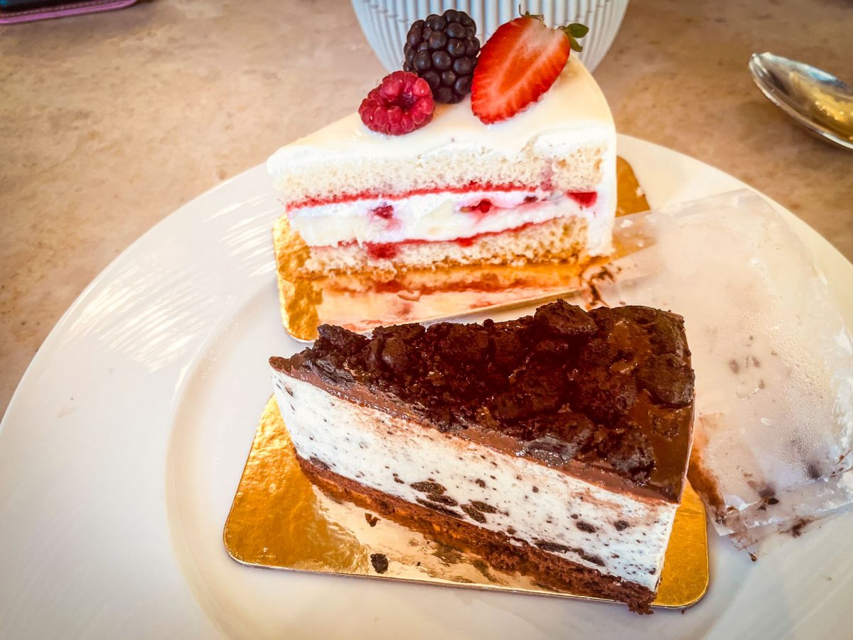 Two slices of dessert on a white plate, including a berry layer cake topped with fresh fruit and a chocolate cookies-and-cream cheesecake.