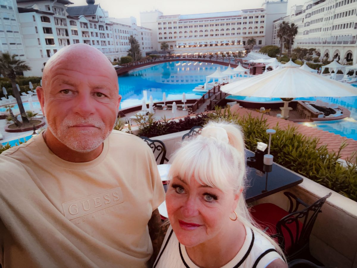 A couple standing on a balcony overlooking a large illuminated swimming pool at a luxury resort in Antalya.