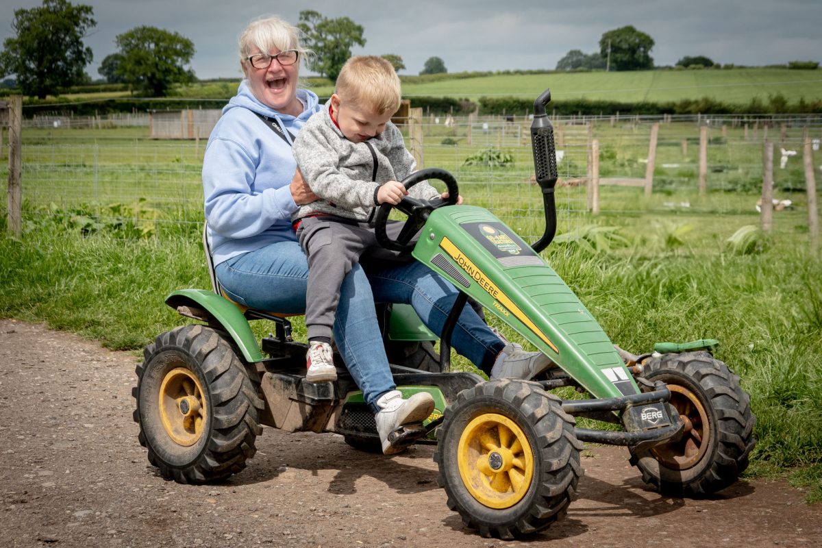 A woman and a young boy riding a green John Deere-branded pedal tractor on a dirt path in a farm field.