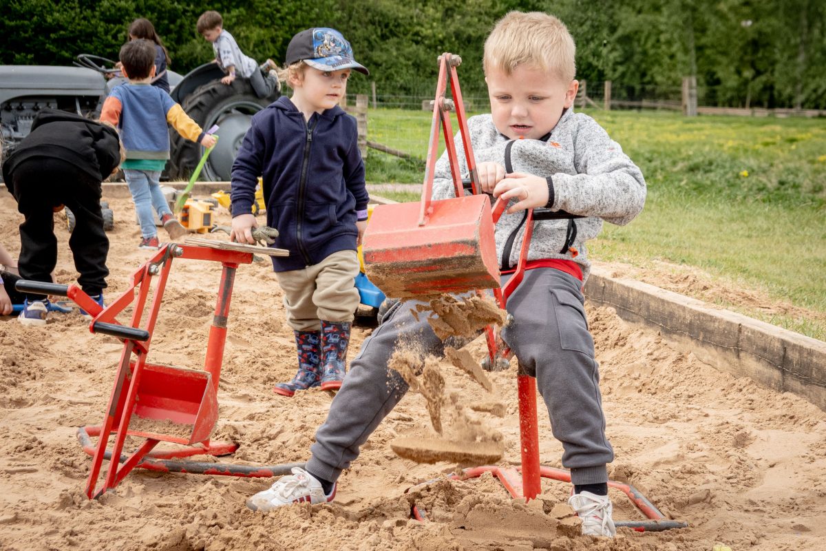 A young boy intensely operating a red, toy excavator in a sandbox, lifting sand. Other children play in the background.