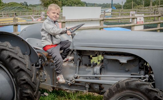 A young boy sitting on the seat of an old grey vintage tractor, pretending to drive.