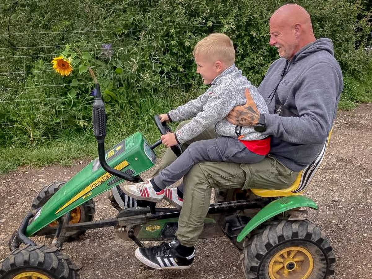 A man with a tattoo and a young boy riding a green John Deere-branded pedal tractor on a dirt path.