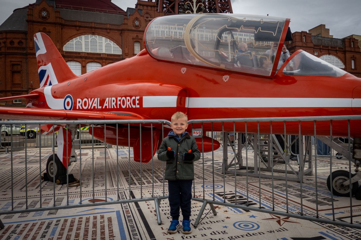 A young boy in a green jacket standing in front of a full-size, bright red Royal Air Force Red Arrows jet on static display, with the Blackpool Tower structure in the background.
