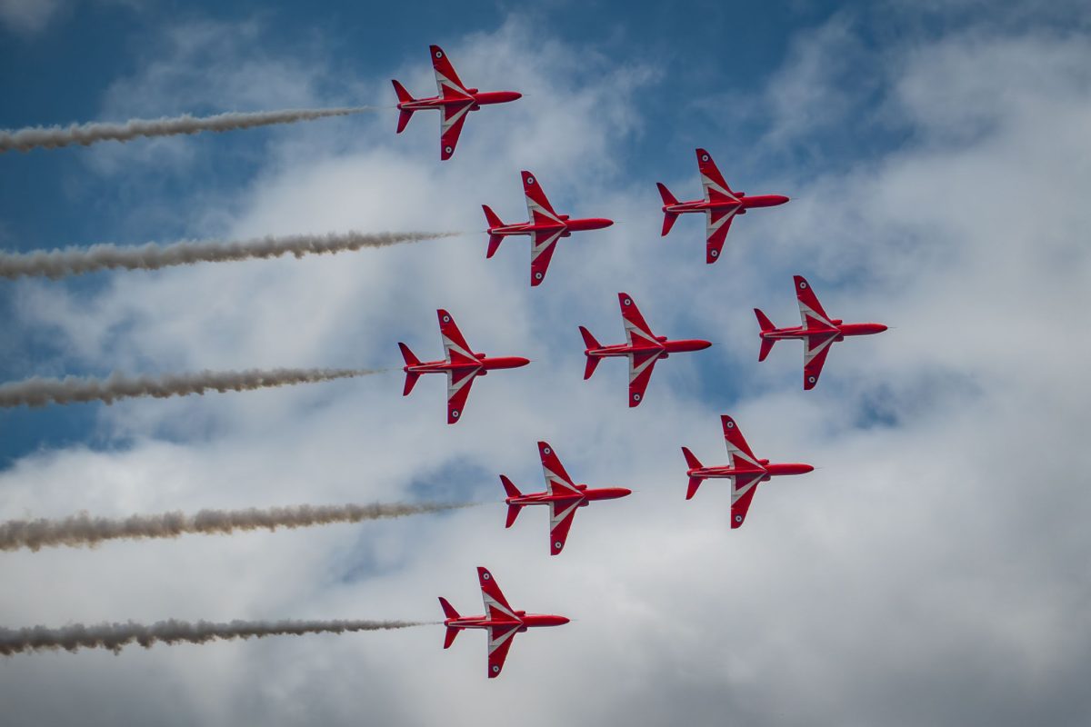 Nine Red Arrows jets flying in a perfect diamond shape formation, with white smoke trails, against a mostly blue sky with scattered white clouds.