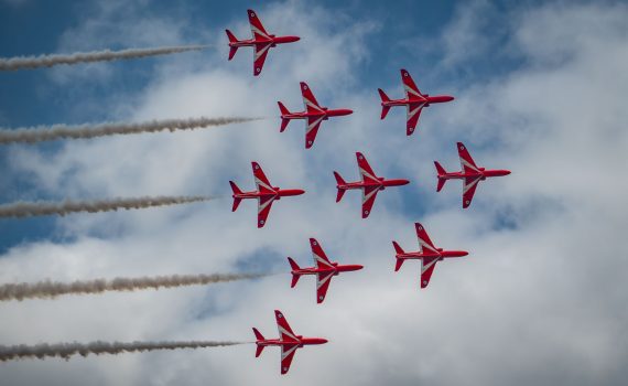Nine Red Arrows jets flying in a perfect diamond shape formation, with white smoke trails, against a mostly blue sky with scattered white clouds.