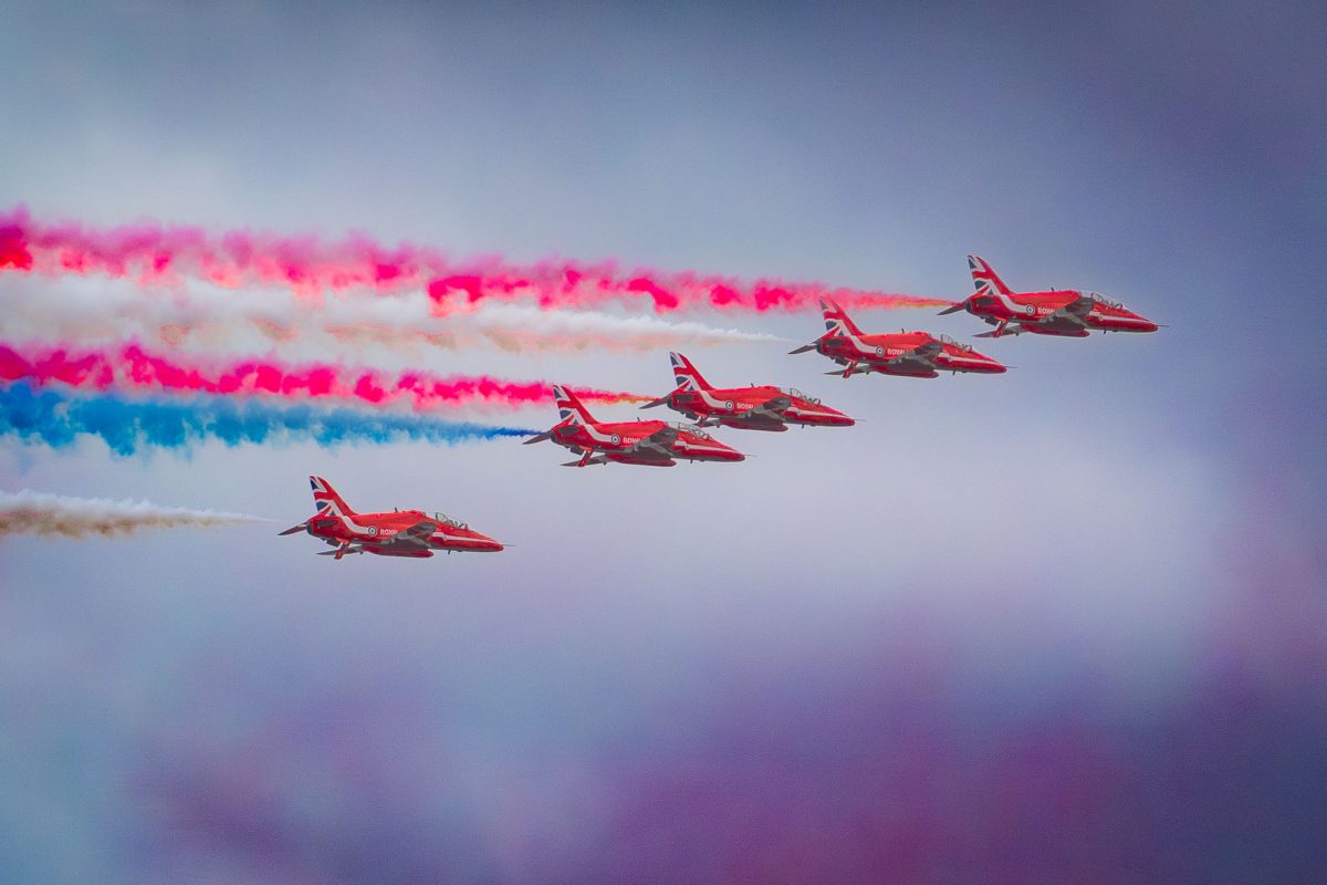 Five Red Arrows jets flying low in a echelon formation, trailing a long, horizontal band of red, white, and blue smoke over a deep purple haze.