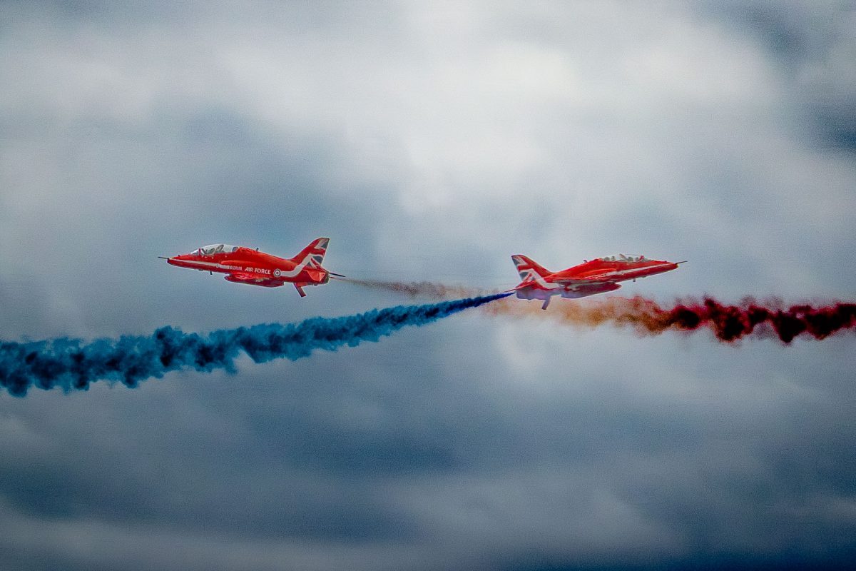 Two Red Arrows jets flying in formation, one emitting blue smoke and the other red smoke, against a dark, cloudy sky.
