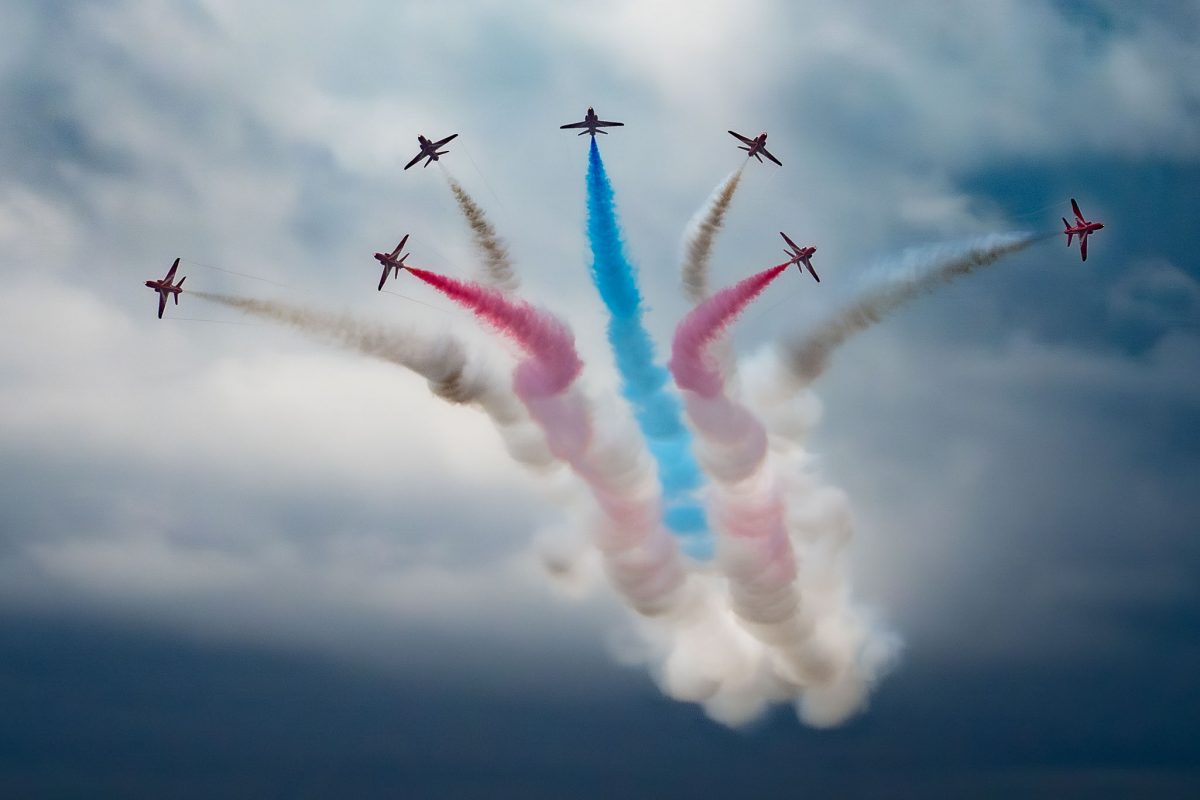 Seven Red Arrows jets breaking outwards from a central point, trailing plumes of red, white, and blue smoke that converge below in a cloud.
