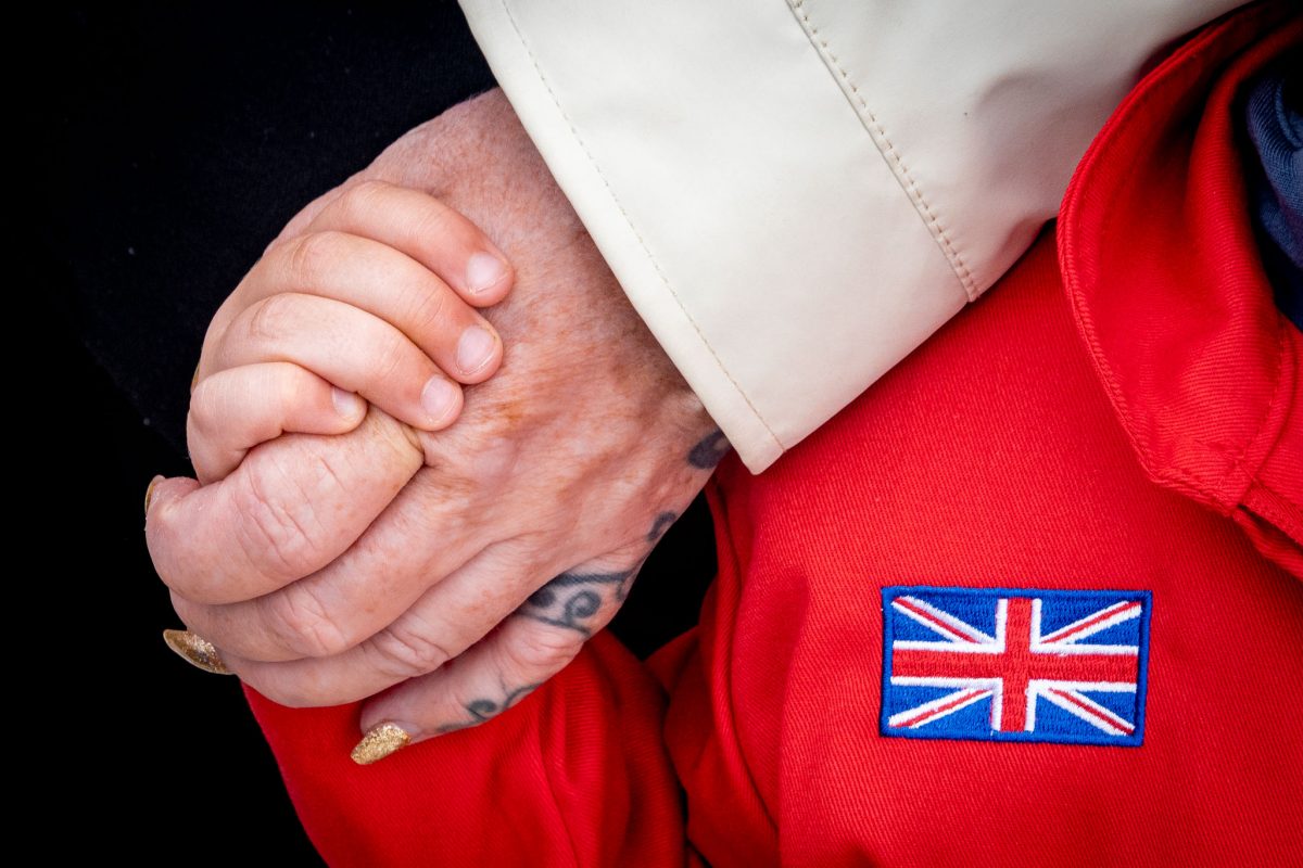 A close-up of an adult's hand gently resting on a child's shoulder, with the child wearing a red suit with a clear Union Jack patch embroidered on it.