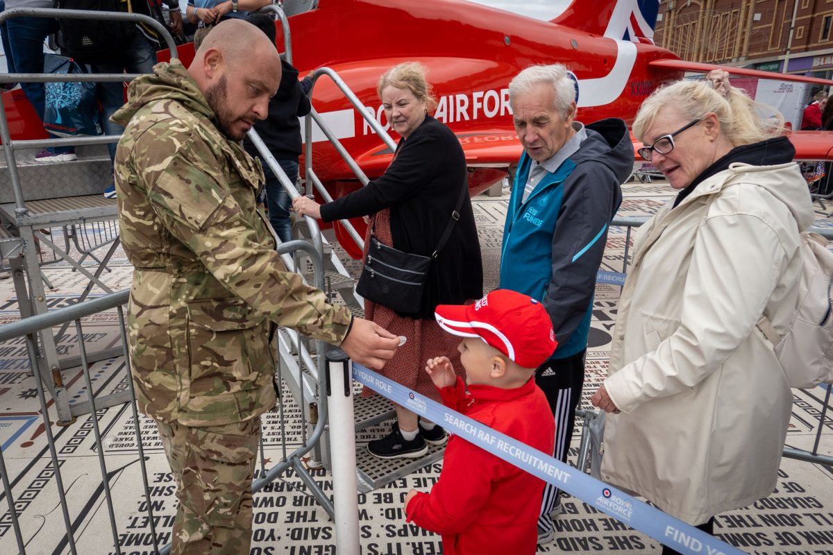 A British soldier in camouflage handing a small item to a young boy in a red cap near a Red Arrows jet display, with a family group watching.