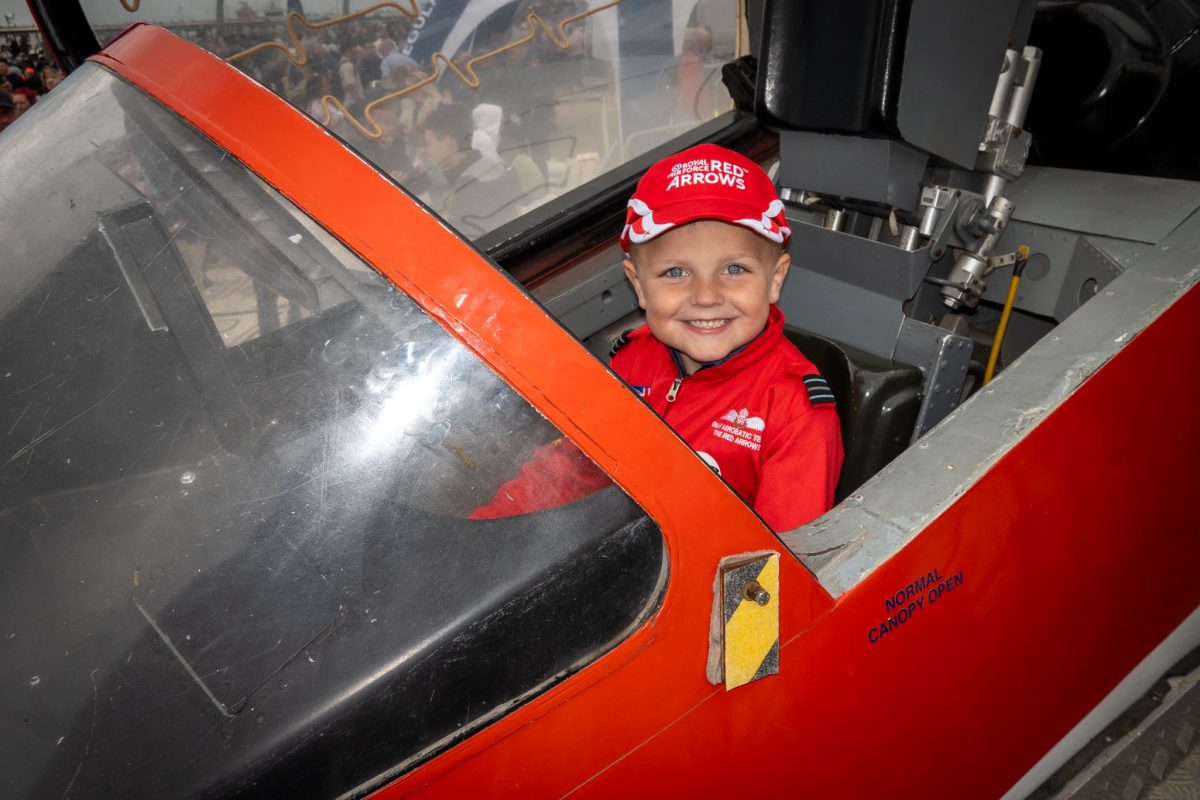 A young boy in a red Red Arrows cap and suit sitting happily in the cockpit of a jet on display, smiling directly at the camera.