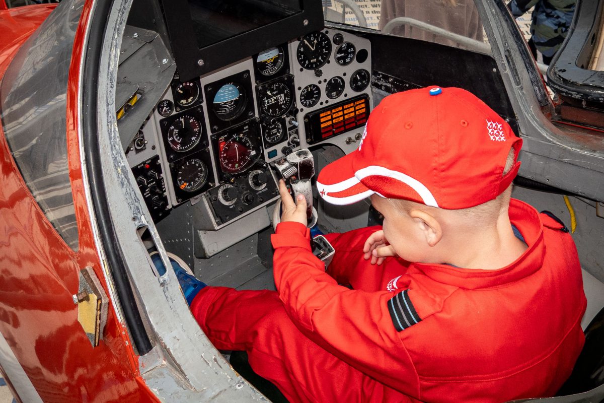 A young boy in a Red Arrows suit and cap sitting in a cockpit and intently examining the detailed instrument panel of the jet.
