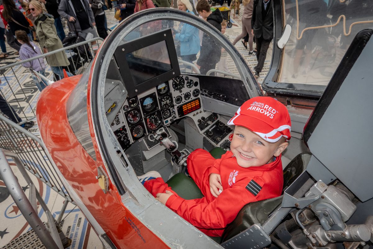 A wide view of the cockpit with a young boy sitting inside, smiling, surrounded by the jet's control panel and a crowd in the background.