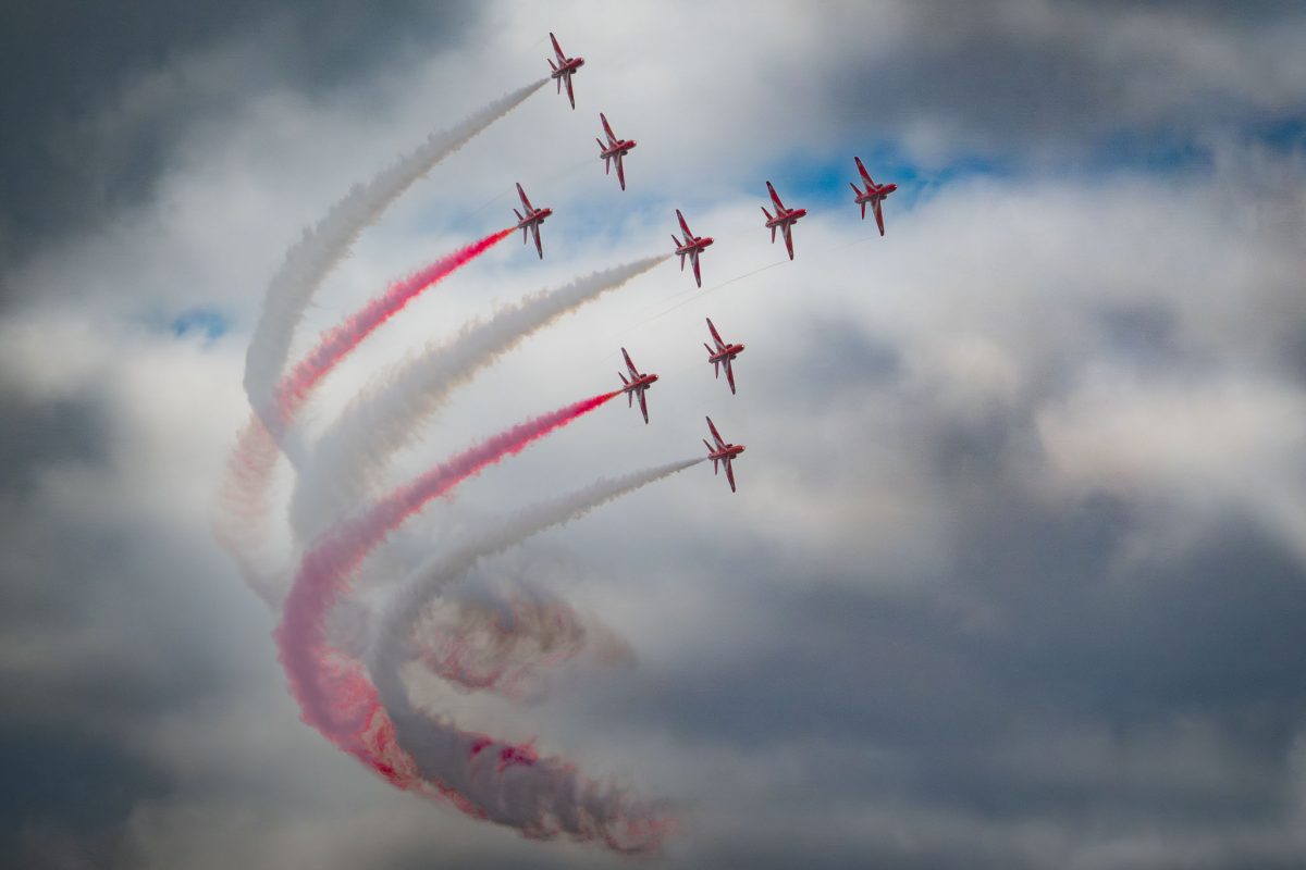 Nine Red Arrows jets ascending in a tight, curved formation through a cloudy sky, leaving white and red smoke trails that form a sweeping arc.