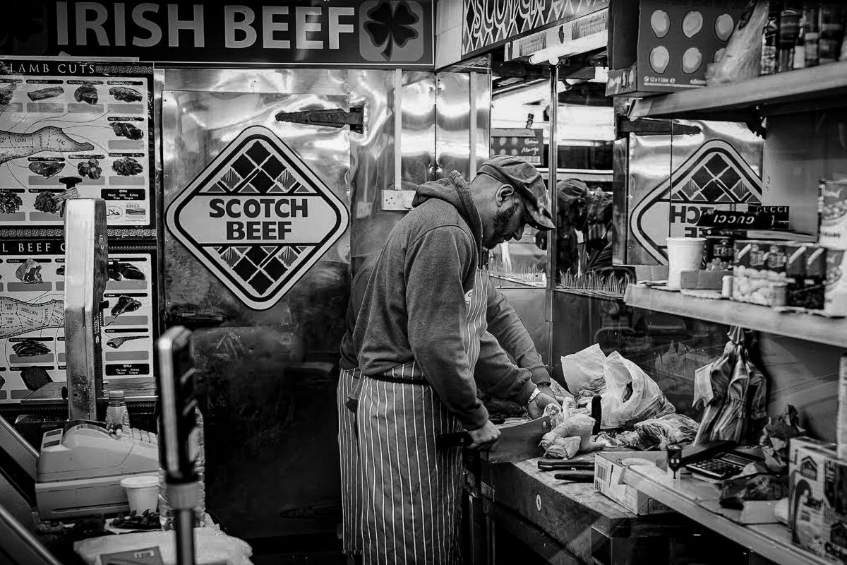 Black and white photo of a butcher in an apron and cap preparing meat behind a counter at a market stall with "IRISH BEEF" and "SCOTCH BEEF" signs.