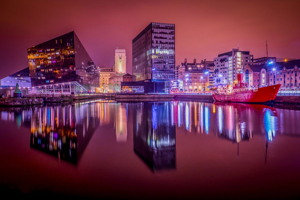 Night photo of Liverpool skyline reflecting in Canning Dock water, showing the modern Museum of Liverpool and the iconic Liver Building tower.
