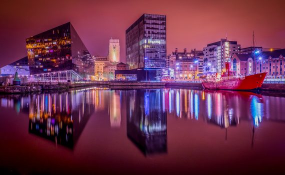 Night photo of Liverpool skyline reflecting in Canning Dock water, showing the modern Museum of Liverpool and the iconic Liver Building tower.