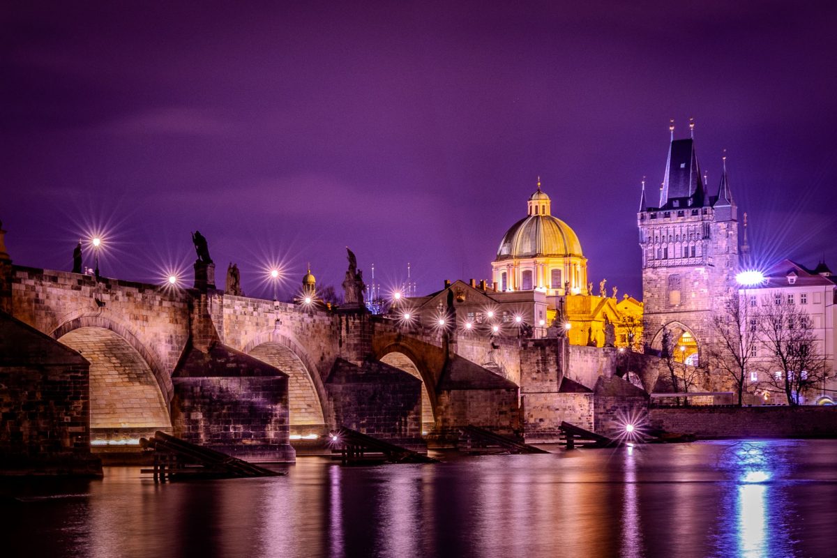 Night photo of the Charles Bridge arches and Baroque statues over the Vltava River, with the brightly lit St. Francis of Assisi Church dome and the Gothic Old Town Bridge Tower in the background, set against a dramatic purple sky.