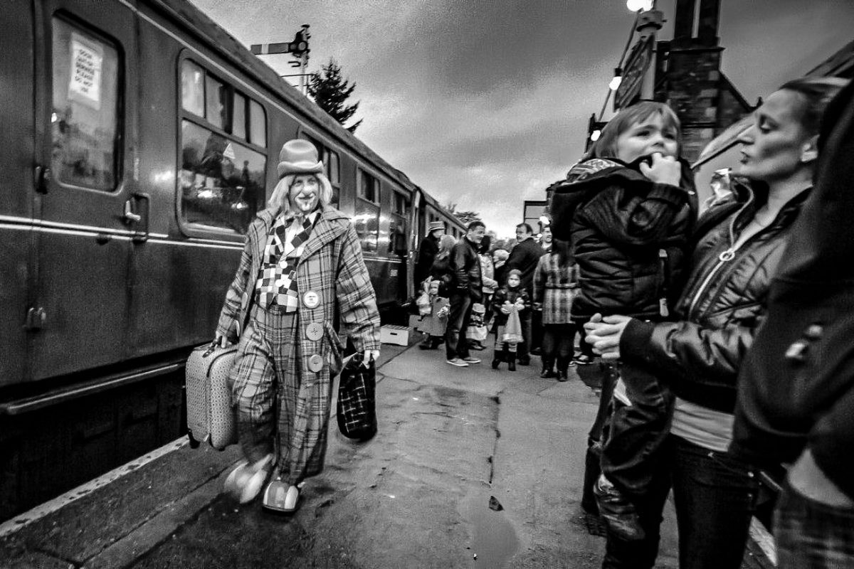 Black and white photo of a clown in a checkered suit, holding a suitcase, walking along a railway platform next to a train, observed by a woman holding a child.