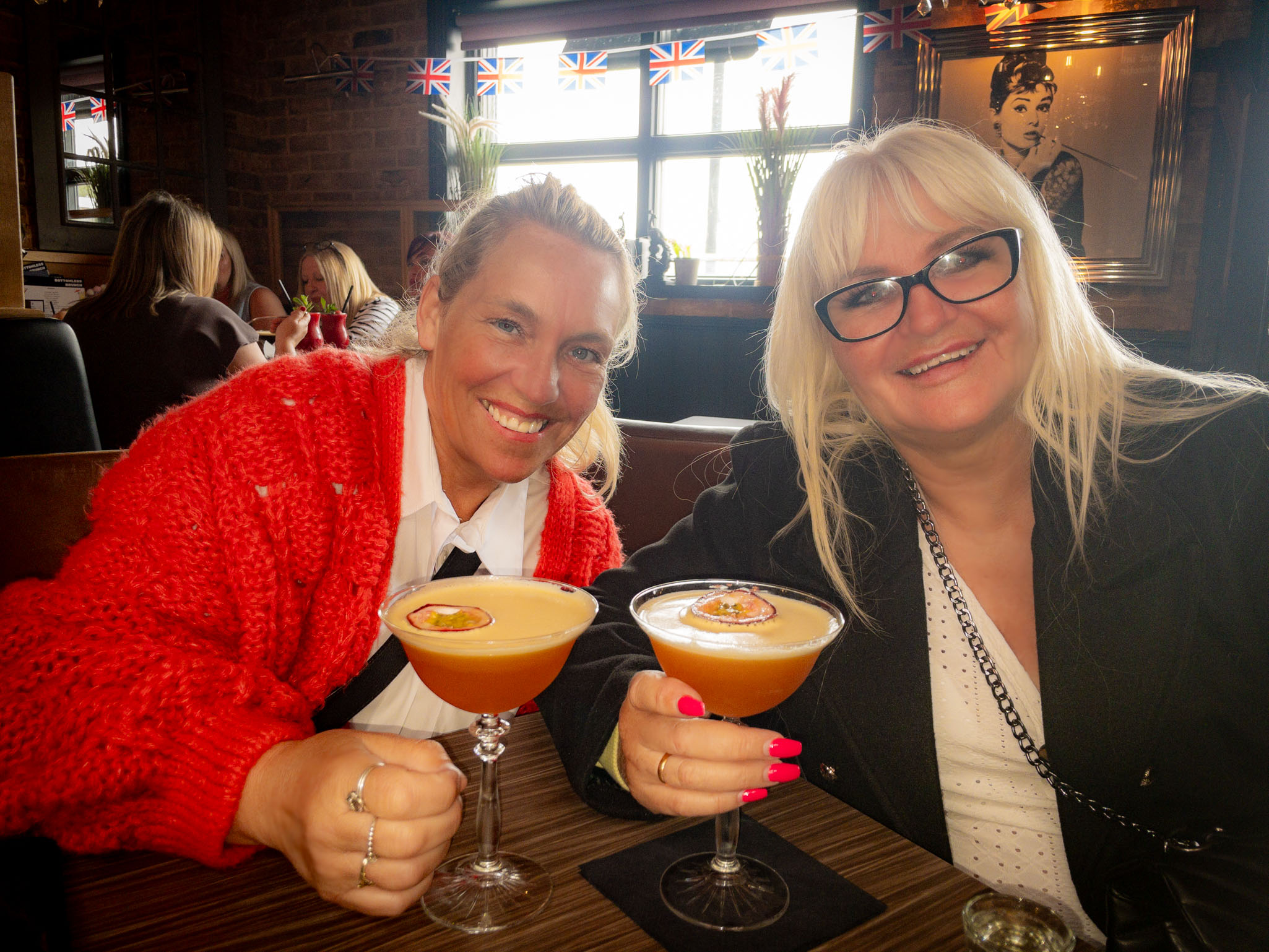 Two smiling women holding passionfruit martini cocktails in a bar with a window view and Union Jack flags