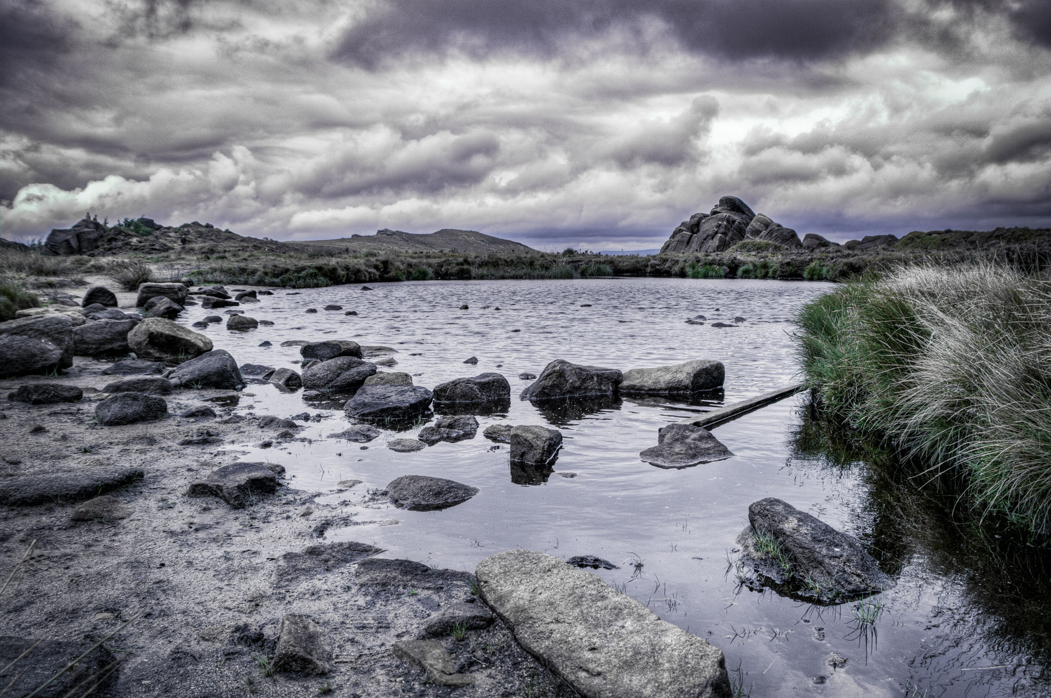 Moody landscape of Doxey Pool at The Roaches in the Peak District, featuring dark water, scattered rocks and stormy skies.