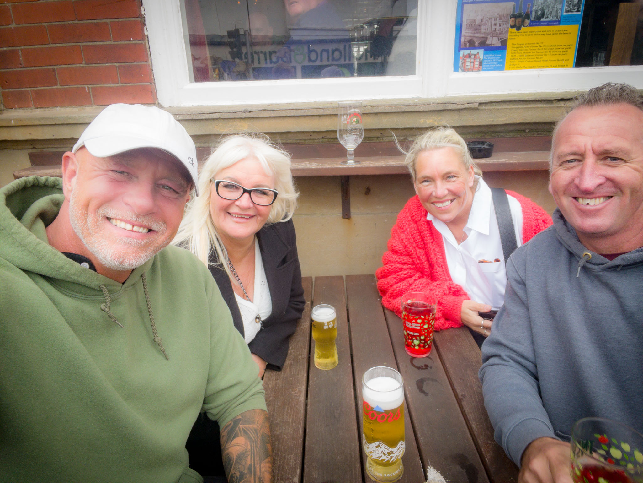 Four smiling friends sitting at a wooden table outdoors, enjoying beers and a red drink at The Dolphin Hotel pub in Whitby, UK.