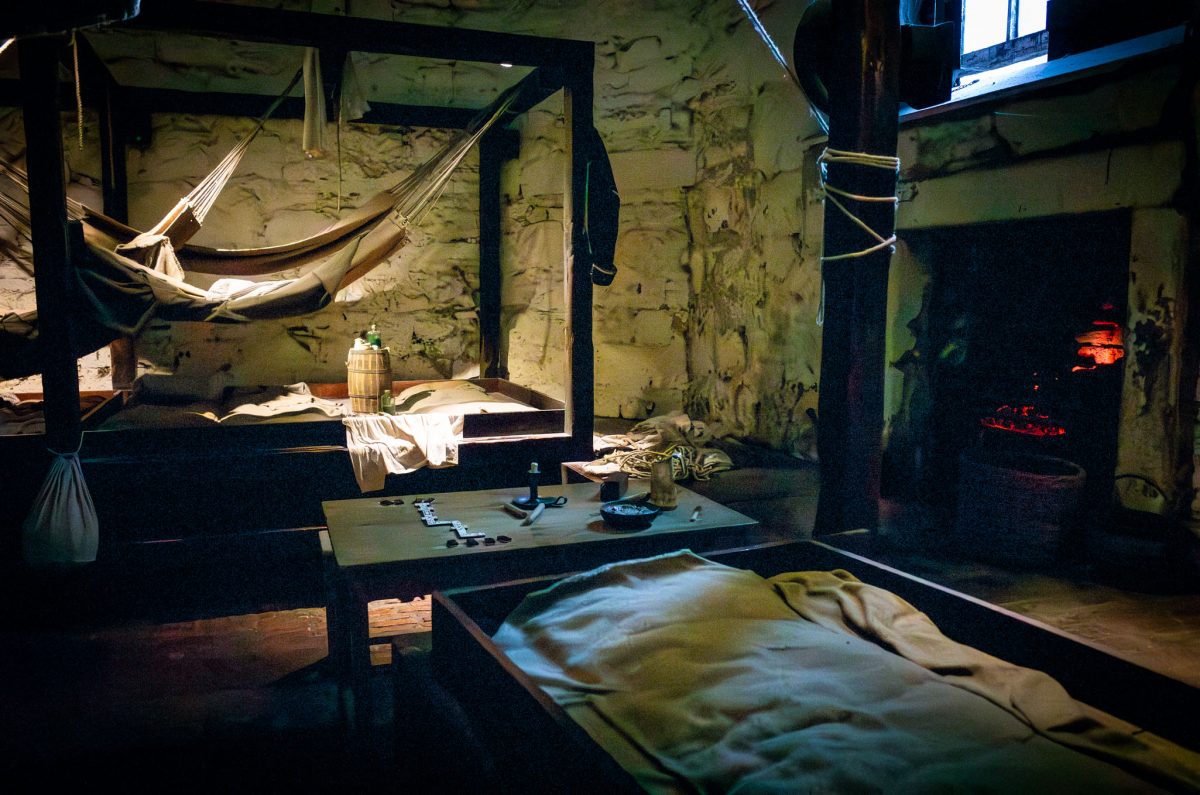A dimly lit, rustic interior scene showing a wooden table, hammocks hanging from the ceiling, and a fireplace, depicting the Castle's former prison life.