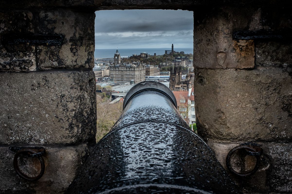 View of the Edinburgh skyline and city buildings through a gun embrasure, with the large black cannon barrel in the foreground.