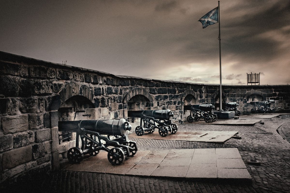 Cannons lined up on the ramparts of Edinburgh Castle under a dramatic, cloudy sky, with the Saltire (Scottish flag) flying high.