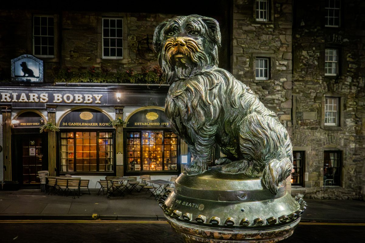 Bronze statue of Greyfriars Bobby sitting atop a water fountain base in front of the Greyfriars Bobby Pub at night in Edinburgh.