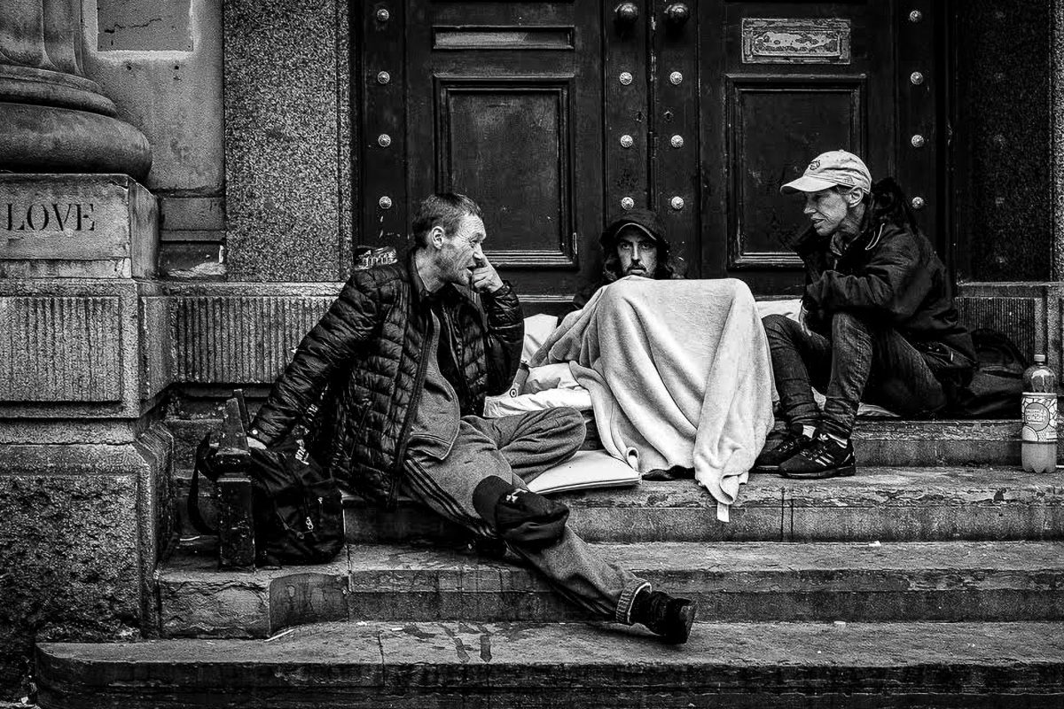 Black and white photo of three adults sitting on stone steps in a large doorway, one man speaking while another two are covered by a blanket. A stone carving with "LOVE" is visible on the left.
