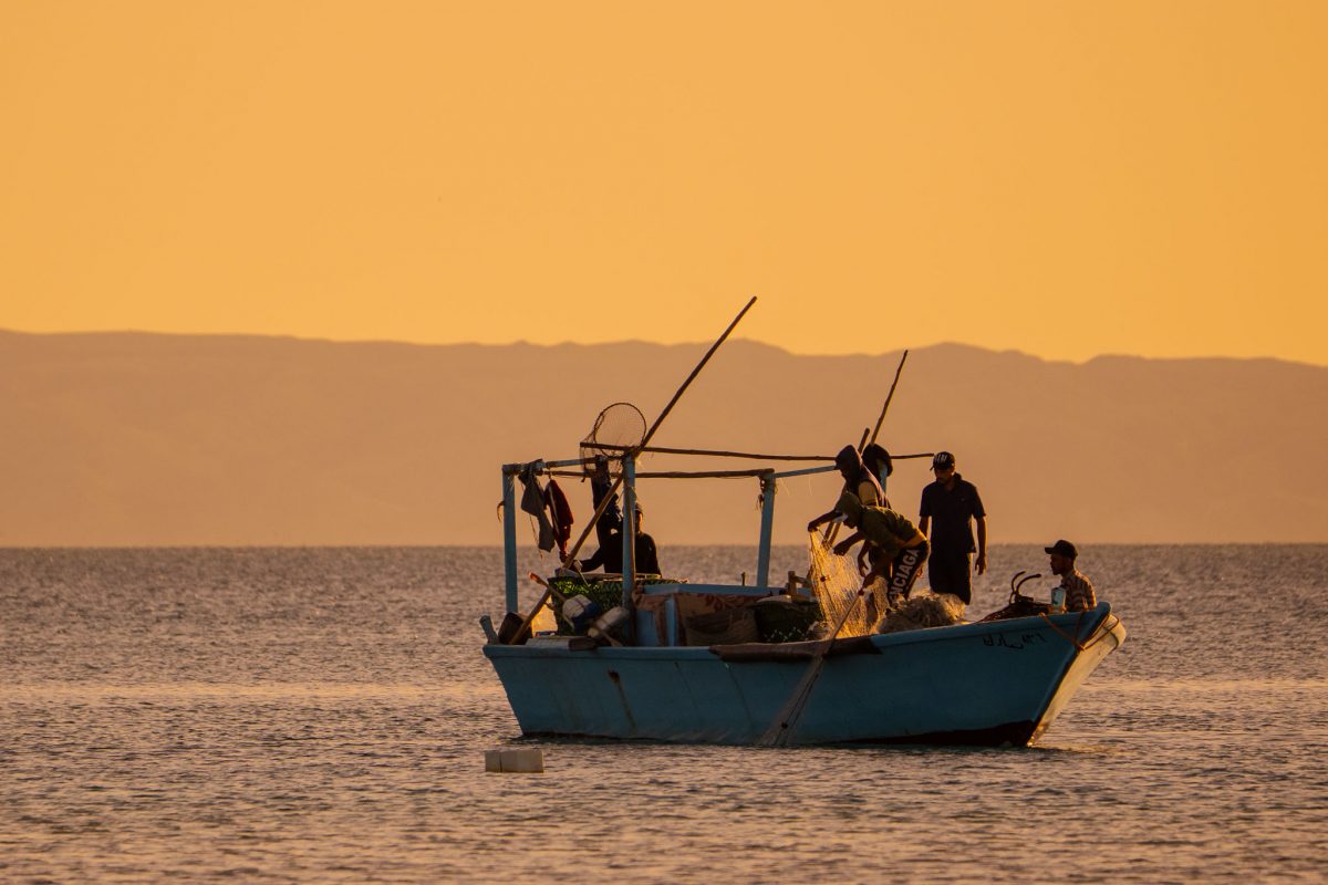 A small blue fishing boat with several silhouetted fishermen working on a net against a warm, orange and gold sunset sky over the Red Sea in Hurghada, Egypt, with hazy distant mountains.