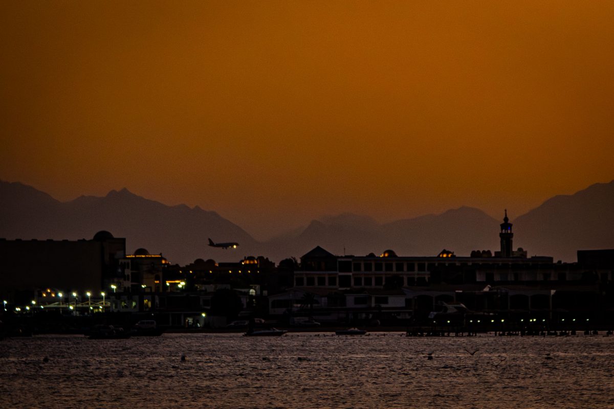 A large Boeing 747 airplane in silhouette making its final approach to Hurghada International Airport, set against a bright orange-red sunset sky with the city's coastal buildings and distant mountains below.