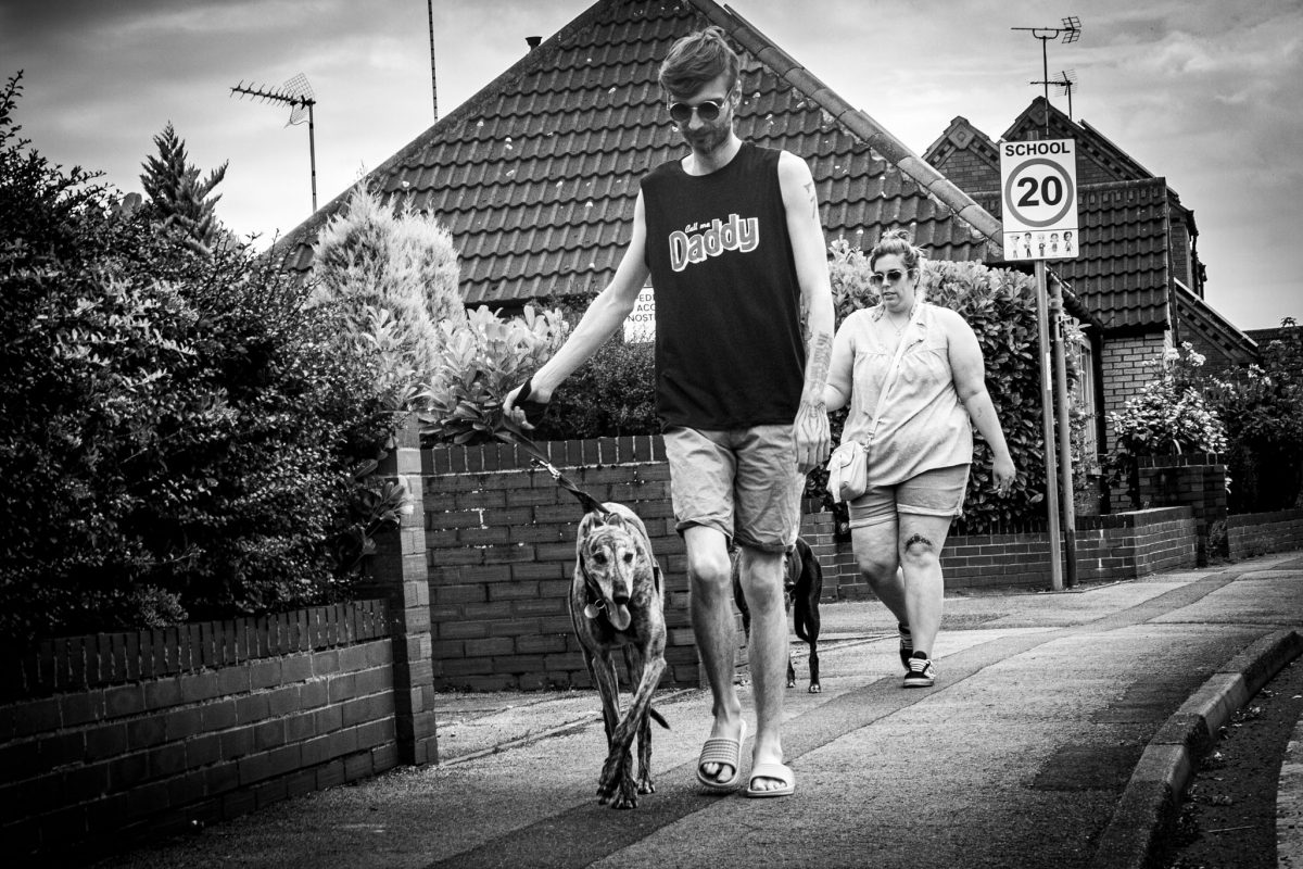 Black and white photo of a man in a "Daddy" vest walking a Greyhound dog on a leash, with a woman and another dog behind him, passing a "School 20" sign in Sheffield.