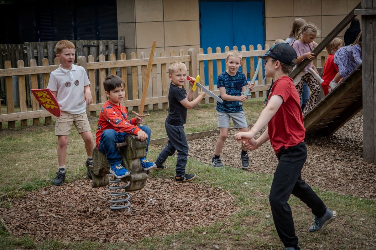 A group of children playing in an outdoor area near a fence, some holding toy swords and shields, acting out a battle or historical scene.