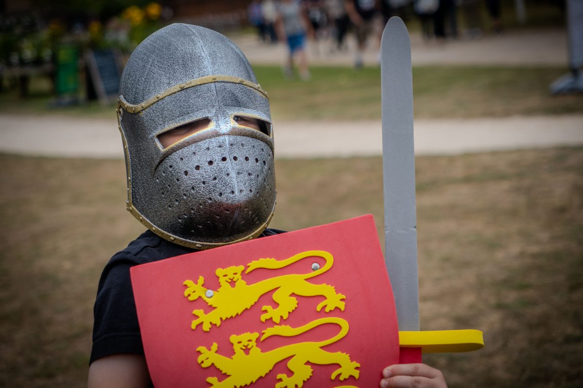 A child wearing a silver medieval helmet with eye slits holds a red shield bearing two yellow lions and a toy sword, focusing on the camera.