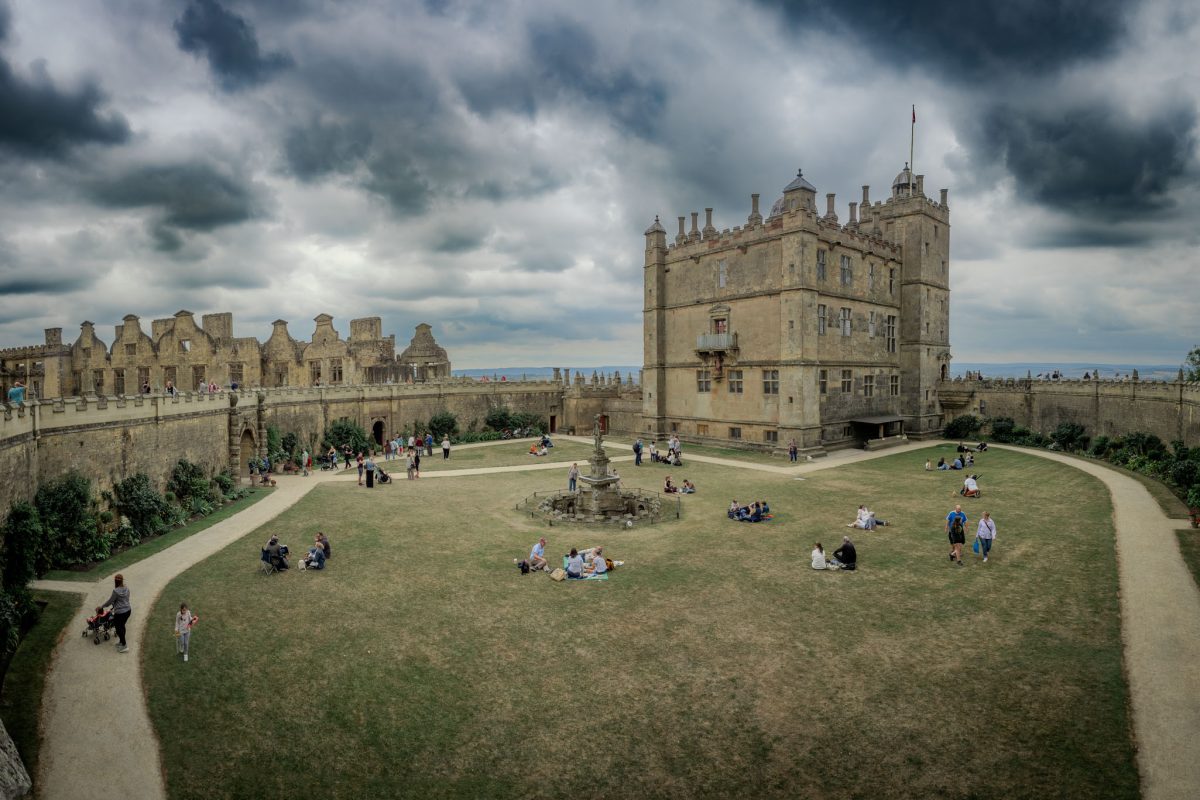 An overview of Bolsover Castle with the main building (the Little Castle) on the right, the ruined Terrace Range on the left, and visitors relaxing on the green courtyard under a dramatic cloudy sky.