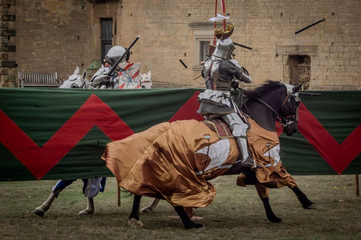 Two knights jousting at full speed; one on a dark horse with gold/orange barding, the other on a light horse. Splintered lances fly in the air above the red and green barrier.