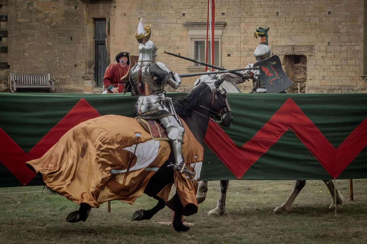 Two knights jousting across the barrier; one knight is on a dark horse with a gold-colored caparison, narrowly avoiding the lance of the knight on a white horse, as an observer looks on.