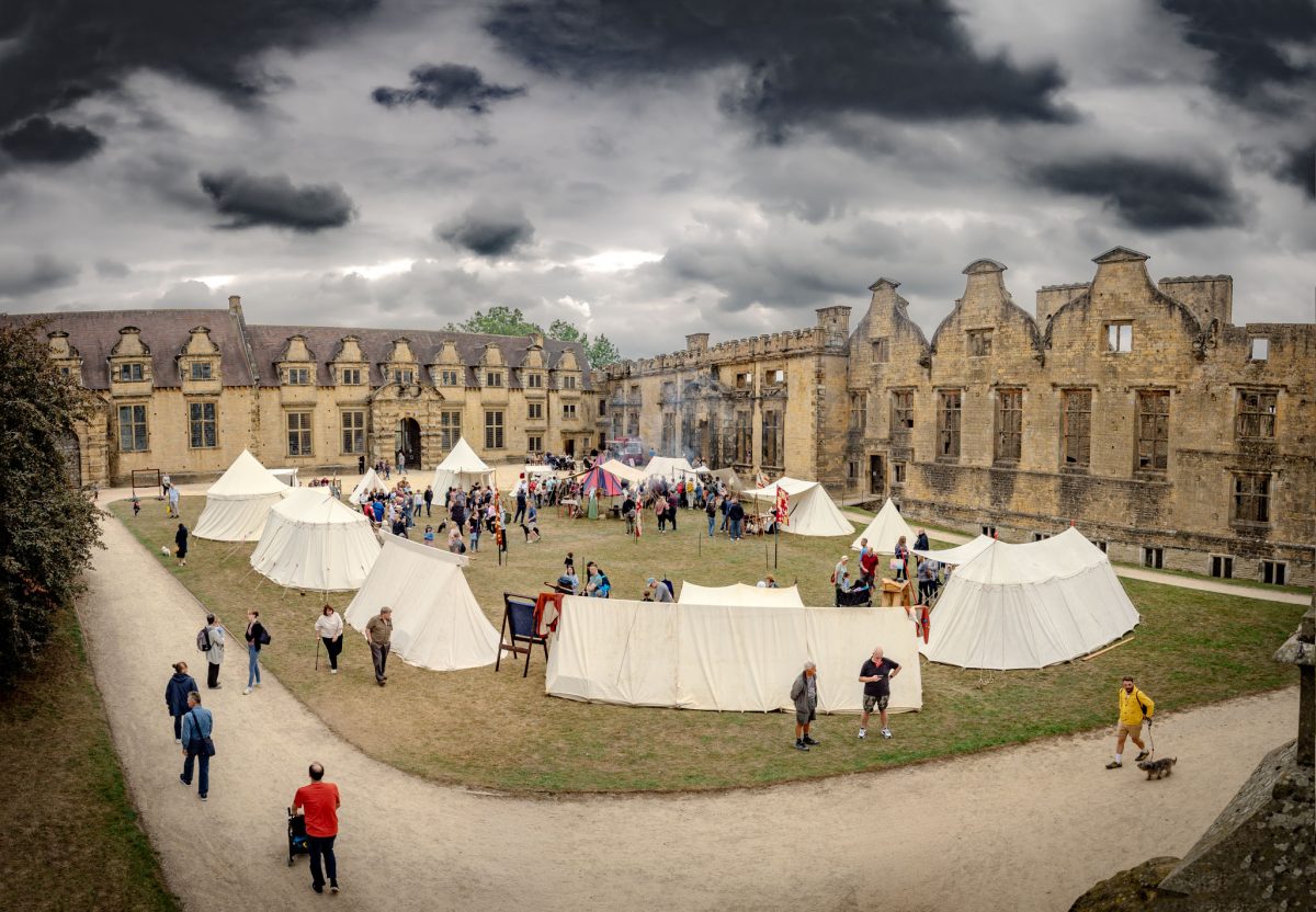 A wide view of the outer bailey of Bolsover Castle, showing the ruined buildings, a crowd of visitors, and a large medieval re-enactment camp with several white conical tents.