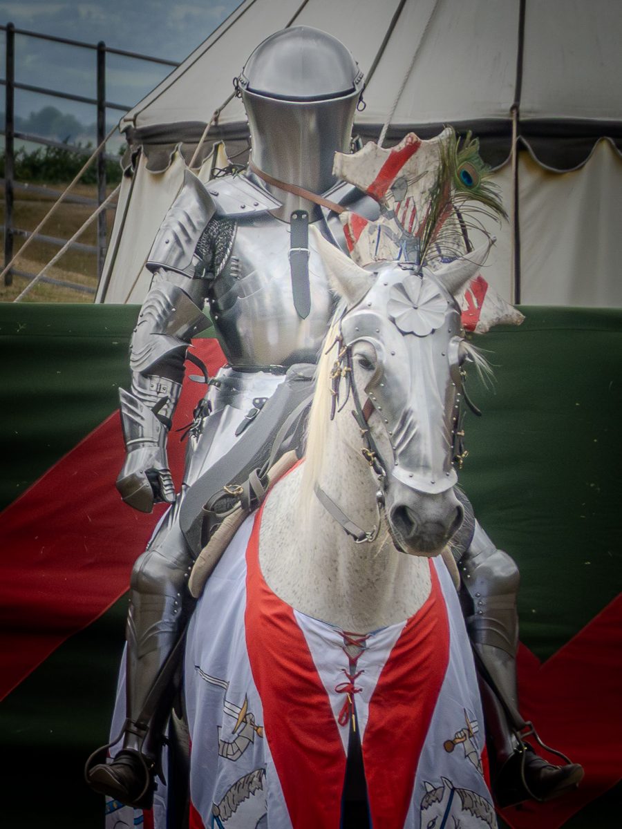 A close-up of a knight in polished plate armor seated on a white, spotted horse, whose barding is white and red, framed by a re-enactment tent and a peacock feather on the helmet.